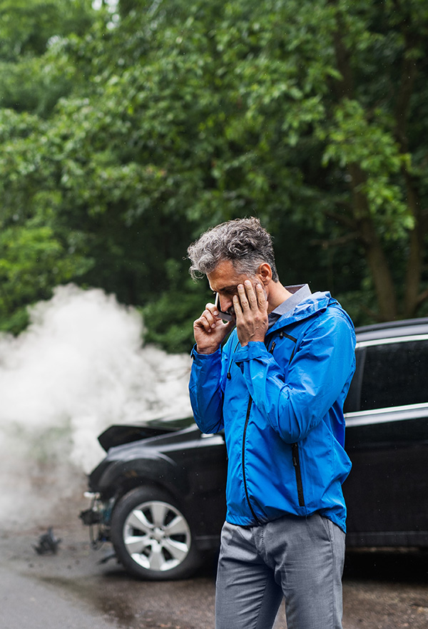Mature man making a phone call after a car accident, smoke in the background.